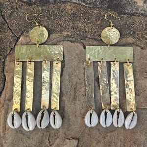 Può includere: Un paio di orecchini color oro con una texture martellata. Ogni orecchino presenta una parte superiore circolare, una sezione centrale rettangolare, barre verticali pendenti e conchiglie di ciprea bianche nella parte inferiore. Orecchini forati.