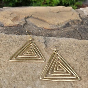 May include: A pair of gold-colored, geometric earrings. Each earring features a triangular frame with a spiral design inside. The earrings have a hook closure and are displayed on a textured, light brown surface.