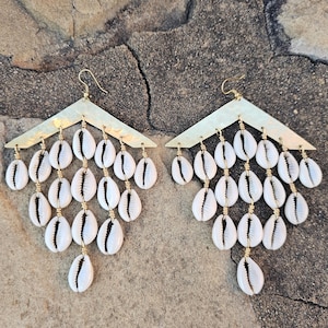 Op de afbeelding: Een paar gouden oorbellen met een driehoekige bovenkant en een cascade-ontwerp van witte kauri-schelpen.