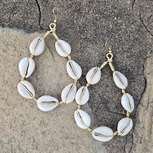 May include: A pair of gold hoop earrings with white seashells strung on the wire. The earrings are dangling and the shells are facing outwards.