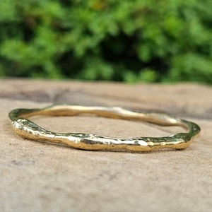 May include: A gold-colored ring with a textured, organic design. The ring is circular and appears to be made of metal. The ring is displayed on a wooden surface with a blurred green background.