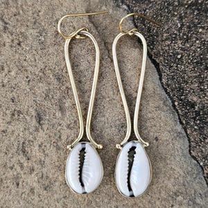 May include: A pair of gold-colored drop earrings featuring a white and black cowrie shell design. The earrings have a long, slender, curved gold wire that connects to the shell. The shells have a natural, organic shape.