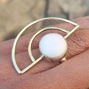 May include: A gold-colored ring with a white, round stone. The ring has a unique design with a semi-circular shape and a circular element around the stone. The ring is on a person's finger.