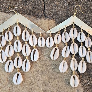 Op de afbeelding: Een paar gouden oorbellen met een driehoekige bovenkant en een cascade van witte kauri-schelpen.