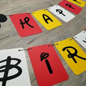 Puede incluir: Una pancarta en blanco y negro con las palabras "Happy Birthday" en letras negras sobre un fondo rojo, blanco y amarillo. La pancarta est&aacute; enhebrada con una cuerda negra.