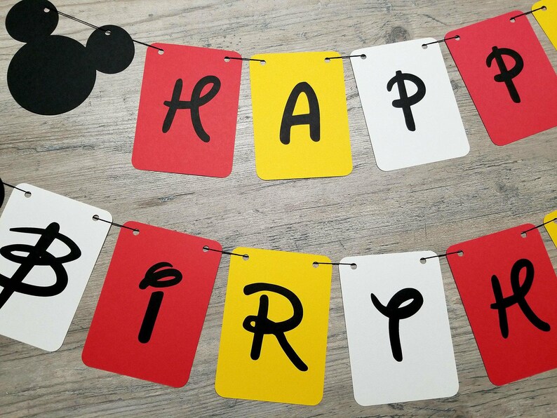 Puede incluir: Una pancarta roja, amarilla y blanca con letras negras que dice "Happy Birthday". La pancarta est&aacute; unida con una cuerda negra y tiene una cabeza de Mickey Mouse negra al principio de la pancarta.