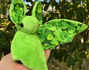 May include: A plush green bat with large ears and wings. The wings are decorated with a pattern of green clovers. The bat is held in a hand against a background of green foliage.