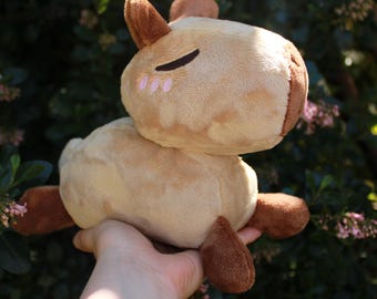 May include: A plush capybara toy featuring a light tan body, dark brown ears and feet, and closed eyes with small pink blush marks on the cheeks. The stuffed animal has a rounded head and a soft, textured surface, presented in a side profile view.
