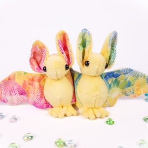 May include: Two plush bat toys with soft yellow bodies and black eyes. Their large, upright ears and wide, outstretched wings feature a vibrant, multicolored fabric pattern with shades of pink, orange, yellow, green, and blue, accented by white floral details.