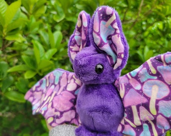 May include: A plush purple bat with large ears and wings. The bat's wings and the inside of its ears feature a colorful pattern of mushrooms and celestial elements. The bat has large, black, round eyes and a soft, fuzzy texture.