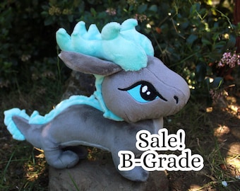 May include: A plush dragon toy featuring a grey body with a bright turquoise mane and tail fin. The creature has large, embroidered blue eyes and a smooth, soft texture. The text "Sale! B-Grade" is printed in white on the lower right side.