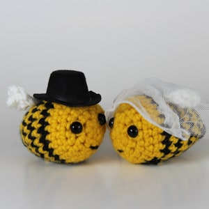 May include: Two crocheted bee plushes, one wearing a black top hat and the other wearing a white veil, facing each other.