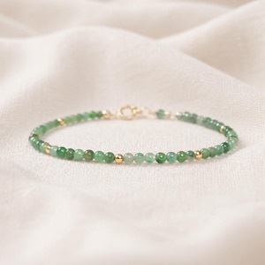 May include: Delicate bracelet featuring small, round, green beads interspersed with gold-colored beads. The bracelet is strung on a thin, gold-colored chain and has a gold clasp. The jewelry is displayed on a white, textured surface.