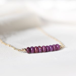May include: A delicate gold chain necklace featuring a row of small, round, deep purple gemstones. The gemstones are evenly spaced and create a linear design. The necklace is displayed on a white, textured surface.