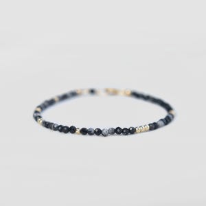May include: A delicate bracelet featuring small, faceted black and gray beads interspersed with gold-colored beads. The bracelet is strung on a thin, flexible cord and has a gold-colored clasp. The jewelry is set against a plain white background.