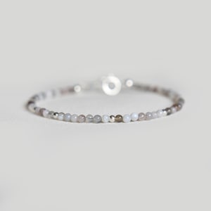 May include: A fine bracelet composed of tiny, spherical beads in grey, brown, and white hues. The beads are strung together with a silver clasp. The bracelet is designed to be worn around the wrist. The overall aesthetic is minimalist and elegant.