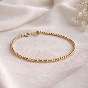 May include: A delicate gold bracelet featuring small, evenly spaced beads. The bracelet has a simple, elegant design with a clasp closure. The jewelry is displayed on a white, textured fabric, creating a soft, neutral background.