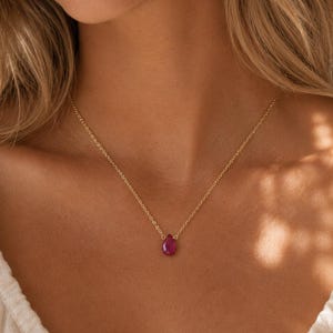 May include: A gold necklace with a teardrop-shaped, deep red gemstone pendant. The delicate chain is visible against the skin, highlighting the pendant's color and shape. The necklace is a simple, elegant piece of jewelry.