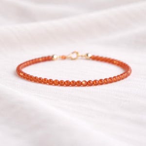 May include: A delicate bracelet featuring small, round, orange beads strung together. The bracelet has a gold-coloured clasp and end pieces. The beads are a warm, reddish-orange colour, and the bracelet is displayed on a white surface.