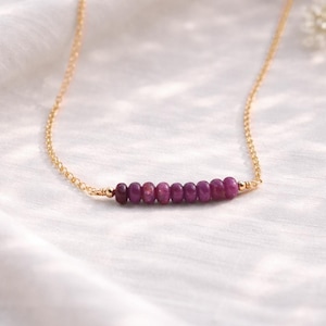 May include: A delicate gold chain necklace featuring a row of small, oval-shaped, deep red gemstones. The gemstones are evenly spaced and set horizontally. The necklace is displayed on a white, textured surface.