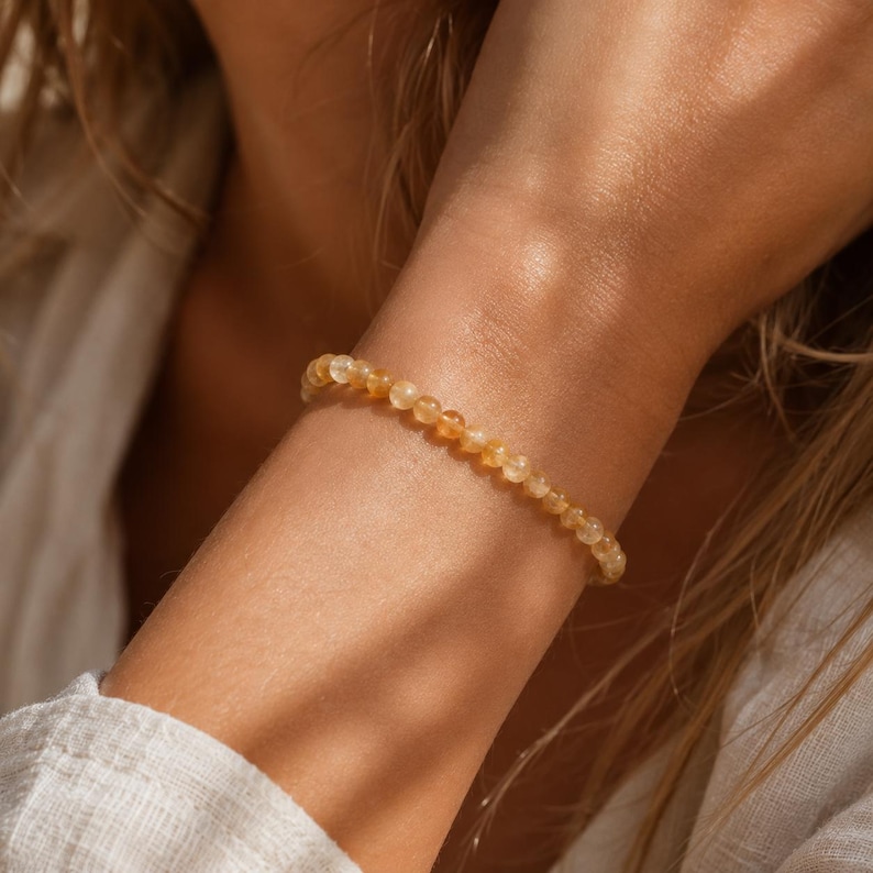 May include: A beaded bracelet featuring small, round, yellow-orange gemstones. The bracelet is worn on a wrist, with the gemstones appearing to be strung together. The bracelet is a warm, golden color, and the beads are uniform in size.