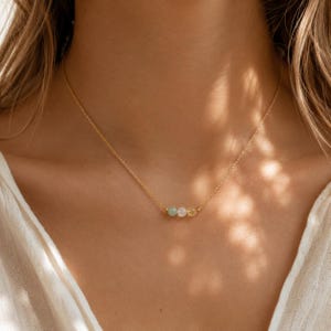 May include: A delicate gold chain necklace featuring three small, round, smooth beads in soft shades of pale green, translucent white, and light yellow, centered along the thin metallic link chain.