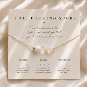 Peut inclure: Un collier à chaîne dorée avec trois perles : une howlite blanche, un quartz transparent et une pierre de lune translucide. Le collier est présenté sur une carte portant le texte THIS FUCKING SUCKS et des descriptions des pierres.