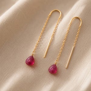 May include: A pair of gold-colored threader earrings with teardrop-shaped, faceted, red gemstones. The earrings hang from a delicate chain and a straight bar. The gemstones are a vibrant shade of red, and the earrings are displayed on a neutral-toned fabric.