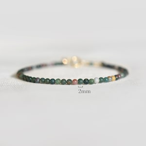 May include: A delicate beaded bracelet featuring small, round, multi-colored stones in shades of dark green, brown, and cream. The beads measure 2mm in diameter as indicated by the text label. A gold-toned metal clasp is visible at the back.