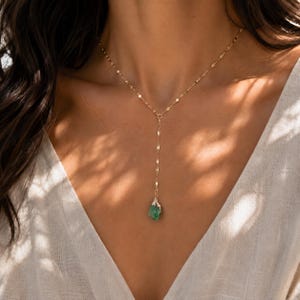 May include: A gold lariat necklace featuring a delicate chain with flat, reflective links and a single raw, uncut green gemstone pendant at the center drop. The necklace rests against a white fabric garment, showcasing a Y-shaped design.