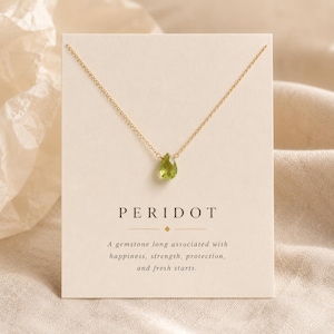 May include: A gold chain necklace featuring a single teardrop-shaped green peridot gemstone pendant. The necklace is displayed on a rectangular white card with the text PERIDOT and a description of the gemstone's associations printed in black.