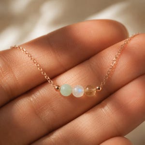 May include: A delicate rose gold chain necklace featuring three small, round, polished beads in light green, iridescent white, and translucent yellow, accented by two tiny rose gold spheres on either side of the central bead arrangement.
