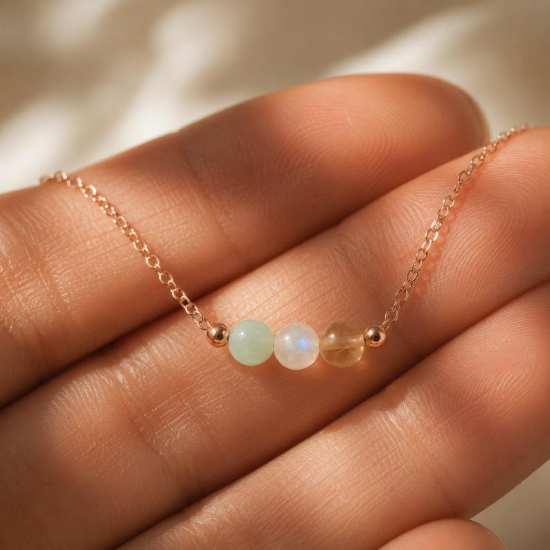 May include: A delicate rose gold chain necklace featuring three small, round, polished beads in light green, iridescent white, and translucent yellow, accented by two tiny rose gold spheres on either side of the central bead arrangement.