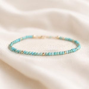 May include: A turquoise beaded bracelet with gold-coloured accents. The bracelet features small, round turquoise beads and gold-coloured beads. The bracelet is displayed on a white, silky fabric.