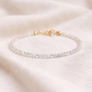 May include: A delicate bracelet composed of small, translucent, faceted beads with an iridescent sheen. The bracelet features a gold-toned metal clasp mechanism at the back, connecting the circular strand of light-reflecting gemstone beads.