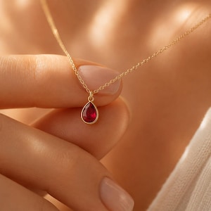 May include: A delicate gold necklace featuring a teardrop-shaped, deep red gemstone pendant. The pendant is set in a gold bezel and hangs from a fine gold chain. The necklace is shown against a soft, neutral background.