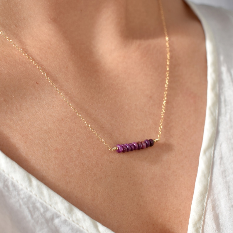 May include: A delicate gold necklace featuring a horizontal bar of small, vibrant purple beads. The necklace is displayed against a white background, highlighting the contrast between the gold chain and the colorful beads.