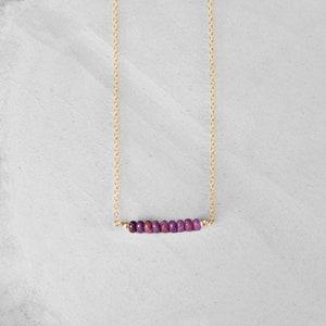 May include: A delicate gold necklace featuring a row of small, vibrant purple beads. The beads are strung horizontally, suspended from a fine gold chain. The necklace is displayed against a neutral, light-colored background.
