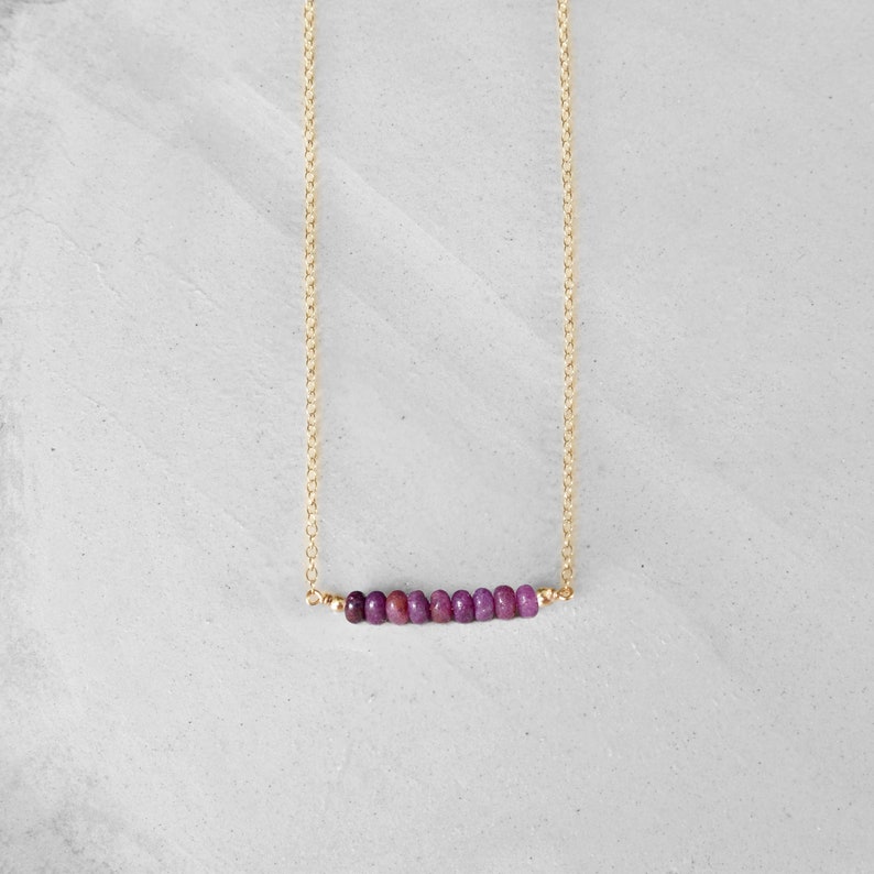 May include: A delicate gold necklace featuring a row of small, vibrant purple beads. The beads are strung horizontally, suspended from a fine gold chain. The necklace is displayed against a neutral, light-colored background.