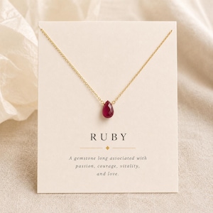 Op de afbeelding: Een gouden ketting met een enkele druppelvormige robijn hanger. De ketting wordt gepresenteerd op een crèmekleurige kaart met de tekst RUBY en een beschrijving van de associaties van de edelsteen met passie, moed, vitaliteit en liefde.