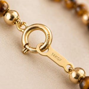 May include: A gold-toned spring ring clasp connected to a flat metal tag, both stamped with 14KGF. The clasp is attached to a strand of polished, spherical tiger's eye beads and a smooth, round gold-toned bead, showcasing a metallic finish.