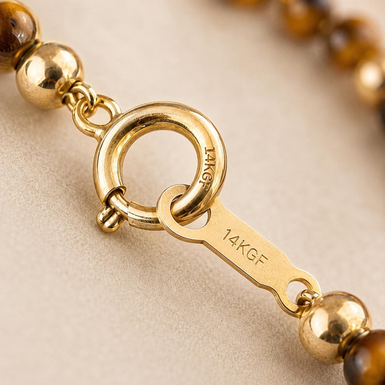 May include: A gold-toned spring ring clasp connected to a flat metal tag, both stamped with 14KGF. The clasp is attached to a strand of polished, spherical tiger's eye beads and a smooth, round gold-toned bead, showcasing a metallic finish.