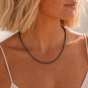 May include: A close-up of a black beaded necklace resting on a person's chest. The necklace is made of small, round beads and is a simple, elegant design. The person is wearing a white top.