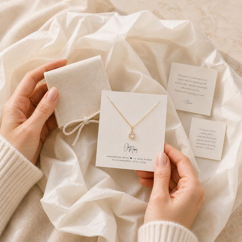 May include: A gold necklace with a teardrop-shaped pendant displayed on a white card featuring the text Olivia Mabrey, Handmade with heart in San Diego, and oliviamabrey.etsy.com, accompanied by a beige velvet pouch and two small note cards.