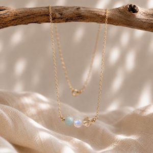 May include: A delicate gold chain necklace featuring three small beads centered at the bottom. The beads consist of a pale blue stone, a translucent white moonstone with a subtle blue sheen, and a clear faceted crystal, accented by tiny gold spheres.
