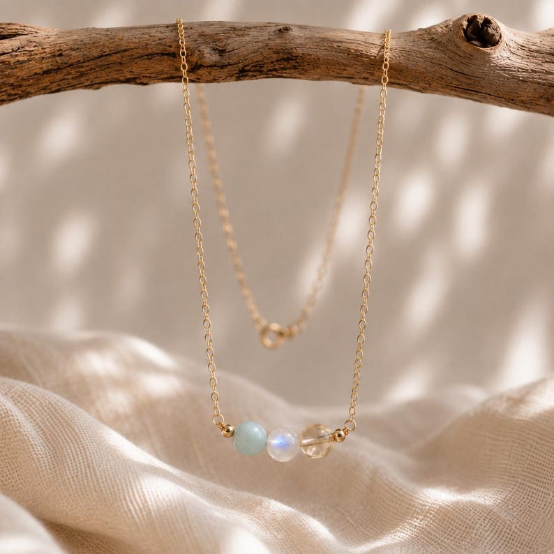 May include: A delicate gold chain necklace featuring three small beads centered at the bottom. The beads consist of a pale blue stone, a translucent white moonstone with a subtle blue sheen, and a clear faceted crystal, accented by tiny gold spheres.