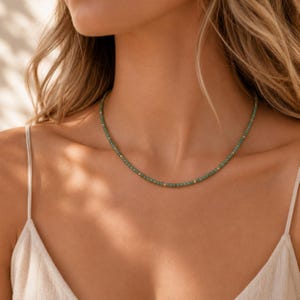 May include: A delicate green beaded necklace with small gold-coloured accents. The necklace is worn around the neck, showcasing its simple yet elegant design. The beads appear to be evenly spaced, creating a refined look.