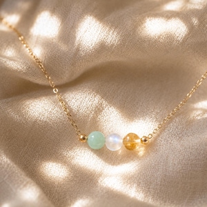 May include: A delicate gold chain necklace featuring three small, round beads in a row: a pale green bead, a translucent white bead, and a golden-yellow bead. Each bead is flanked by tiny gold-toned metal spacers on a thin, polished gold-colored chain.