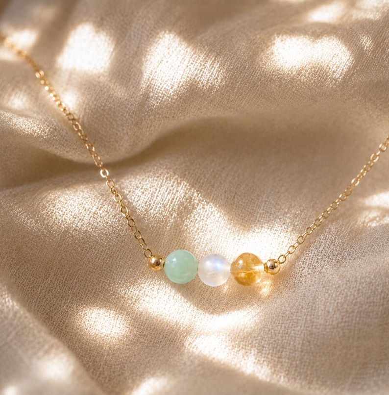 May include: A delicate gold chain necklace featuring three small, round beads in a row: a pale green bead, a translucent white bead, and a golden-yellow bead. Each bead is flanked by tiny gold-toned metal spacers on a thin, polished gold-colored chain.