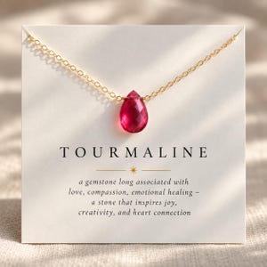 May include: A gold chain necklace featuring a faceted, teardrop-shaped pink tourmaline gemstone pendant. The necklace is displayed on a white card printed with the text TOURMALINE and a description of the gemstone's associations with love.